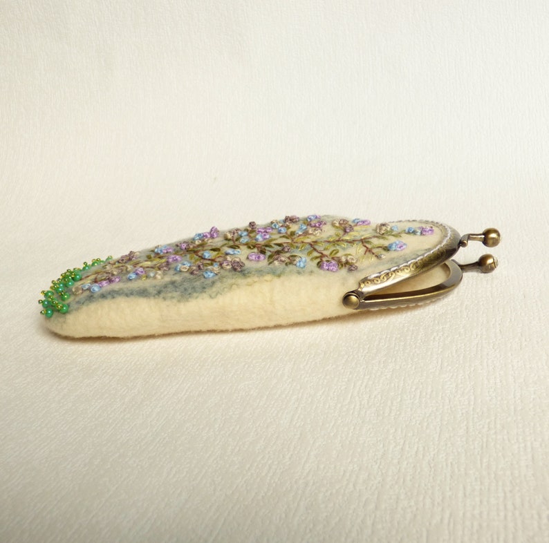 May include: A white felt coin purse with a floral embroidery design in shades of purple, blue, and green. The purse has a brass clasp.