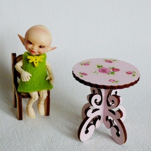 Puede incluir: Una pequeña muñeca vestida de verde con orejas de elfo se sienta en una silla de madera. Una mesa rosa con un diseño floral se encuentra frente a la muñeca.