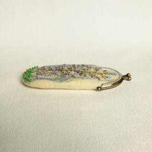 May include: A white felt coin purse with a floral embroidery design in shades of blue, purple, and green. The purse has a brass clasp.