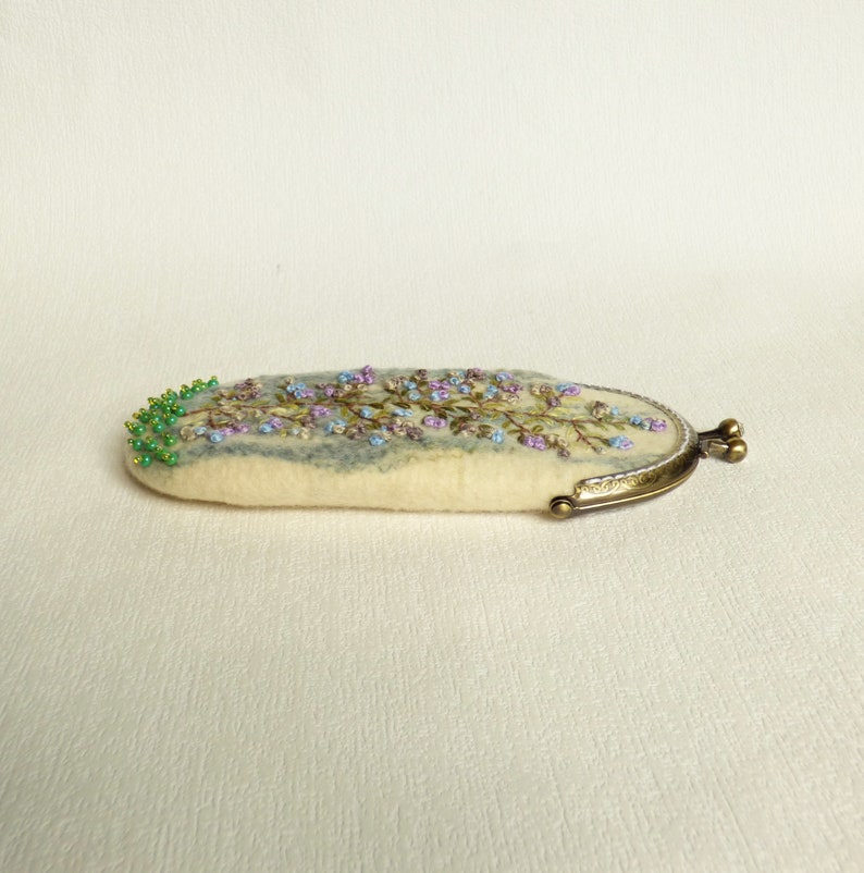 May include: A white felt coin purse with a floral embroidery design in shades of blue, purple, and green. The purse has a brass clasp.