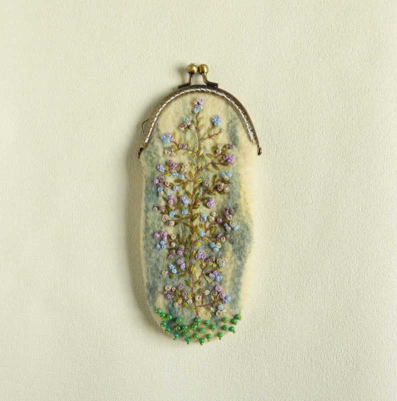 May include: A small, white felt pouch with a floral embroidery design. The pouch has a brass clasp and is decorated with green beads.