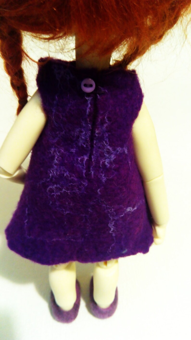 May include: A purple felt dress with a button closure in the back. The dress is worn by a doll with red hair.