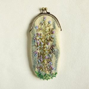 May include: A small, oval-shaped coin purse made of felted wool with a floral embroidery design. The purse has a metal clasp and is decorated with green beads around the bottom.