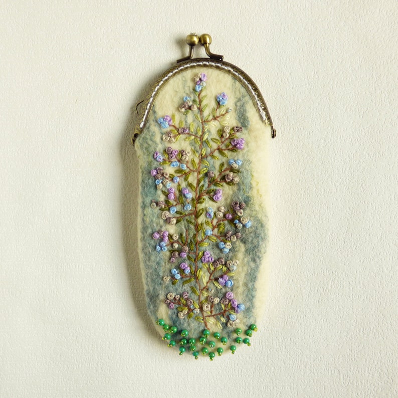 May include: A small, oval-shaped coin purse made of felted wool with a floral embroidery design. The purse has a metal clasp and is decorated with green beads around the bottom.