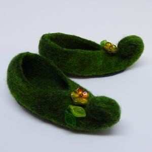 Könnte beinhalten: Ein Paar grüne Filzpantoffeln mit dekorativen Blumenverzierungen. Die Pantoffeln haben eine spitze Zehenpartie und eine abgerundete Ferse.