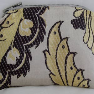 May include: A small, zippered pouch with a beige background and a black and gold floral pattern. The pouch is made of a soft, textured fabric.
