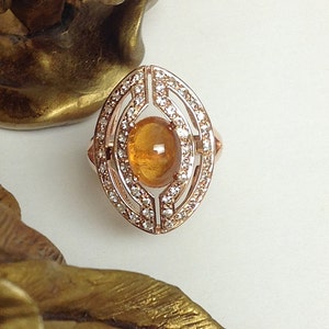 May include: A rose gold ring featuring a large oval-shaped orange gemstone set within a delicate diamond halo.