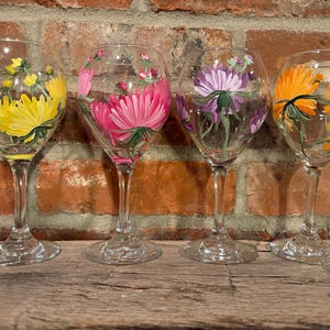 May include: Four clear wine glasses with hand-painted floral designs. Each glass features a different flower: yellow, pink, purple, and orange. The flowers are painted on the glass with green stems and leaves.