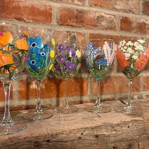 May include: Five clear glass wine glasses, each featuring unique hand-painted floral designs. The glasses display vibrant orange, blue, purple, pink, and red flowers with green stems and leaves painted directly onto the exterior of the glass bowls.
