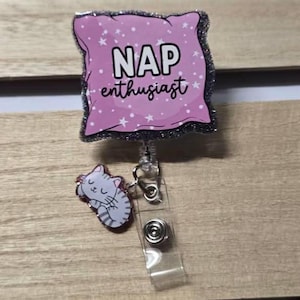 May include: A retractable badge reel with a pink pillow design that reads "NAP enthusiast" in white text. The reel also features a small, sleeping cat charm. The border of the pillow is glittery.