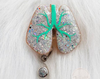 May include: A glittery badge reel in the shape of lungs. The lungs are filled with silver glitter and have a teal-colored bronchial tree design. The badge reel has a silver retractable clip and a round, glittery button.