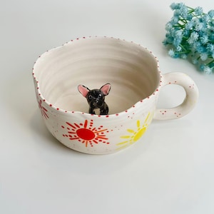 Könnte beinhalten: Eine große, cremefarbene Keramik-Tasse mit Henkel, verziert mit roten und gelben Sonnenmustern. Im Inneren der Tasse befindet sich eine kleine, schwarz-weiße Französische Bulldoggen-Figur aus Keramik. Der Rand der Tasse ist rot gepunktet.