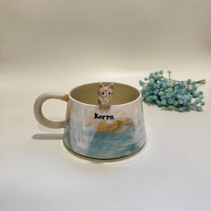 Könnte beinhalten: Eine Keramik-Tasse mit einem bemalten Design in Blau-, Orange- und Weißtönen. Eine kleine, dekorative Katzenfigur sitzt auf dem Rand. Der Name "Korra" steht auf der Seite der Tasse. Ein Zweig blauer Blumen befindet sich im Hintergrund.