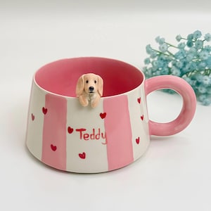 Könnte beinhalten: Ein Keramikbecher mit vertikalen rosa und weißen Streifen, verziert mit kleinen roten Herzen. Der Name Teddy ist in Rot auf der Vorderseite geschrieben. Eine kleine, dreidimensionale hellbraune Hundefigur ist am inneren Rand befestigt und schaut darüber.
