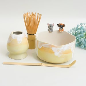 Op de afbeelding: Een matcha theeset met een keramische kom en gardehouder met een geel en wit druipglazuur. De kom heeft twee kleine hondjes op de rand. Een bamboe garde en een lange, gebogen bamboe theeschep zijn in de set inbegrepen.