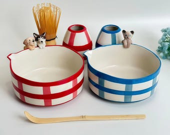 May include: Two ceramic bowls with red and blue plaid patterns. Each bowl has a white interior and is decorated with a small animal figurine. A bamboo whisk and scoop are also present, suggesting a tea set.