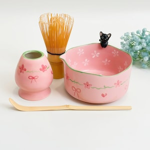 Op de afbeelding: Een roze keramische matchakom met een klein zwart kattenfiguurtje op de rand, een bijpassende roze gardehouder, een bamboe garde en een bamboe lepel. De kom en houder zijn voorzien van handgeschilderde roze kersenbloesems, groene ranken en strikjes.