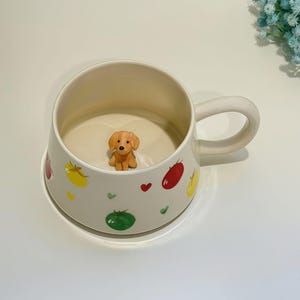 Op de afbeelding: Een crèmekleurige keramische mok met een grote handgreep, versierd met kleurrijke fruitillustraties en een klein, bruin hondenbeeldje aan de binnenkant. De mok heeft een brede opening en een afgeronde basis.