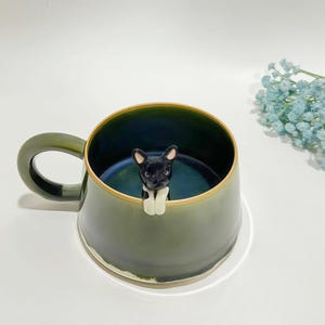 Może przedstawiać: Ceramiczny kubek z gradientowym oliwkowozielonym wykończeniem na zewnątrz i głębokim niebieskim wnętrzem. Mała, rzeźbiona czarno-biała figurka psa jest przymocowana do wewnętrznej krawędzi, z głową i przednimi łapami opartymi o brzeg kubka. Uchwyt jest oliwkowozielony.