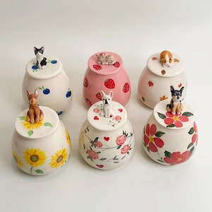 Op de afbeelding: Een verzameling van zes keramische potten met deksels, elk versierd met een ander dierenbeeldje. De potten hebben verschillende ontwerpen, waaronder bosbessen, aardbeien, zonnebloemen, rozen en bloempatronen. De potten zijn wit, roze en crèmekleurig.
