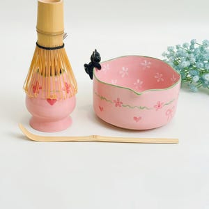 May include: A matcha tea set featuring a pink ceramic whisk holder with red hearts, a bamboo whisk, a bamboo scoop, and a pink ceramic bowl with a green rim, white flowers, and a small black cat figurine perched on the edge of the bowl.