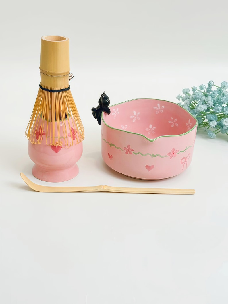 May include: A matcha tea set featuring a pink ceramic whisk holder with red hearts, a bamboo whisk, a bamboo scoop, and a pink ceramic bowl with a green rim, white flowers, and a small black cat figurine perched on the edge of the bowl.