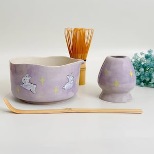 Op de afbeelding: Een lavendelkleurige keramische matchakom en gardehouder met witte konijnillustraties en gele steraccenten, vergezeld door een lichtbruine bamboe matchagarde en een lange, gebogen bamboe theeschep voor het bereiden van matcha.