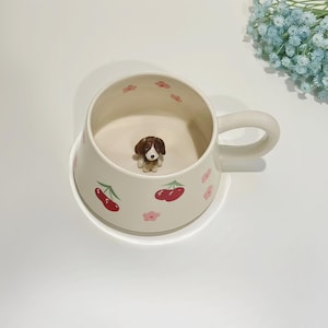 May include: Cream ceramic mug with a handle, decorated with red cherries and pink flowers. A small dog figurine sits inside the mug. The mug rests on a matching saucer.