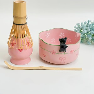 May include: A pink ceramic matcha bowl with a black cat figurine on the rim, a bamboo whisk on a matching pink stand, and a long bamboo scoop. The bowl features painted pink flowers, green vines, and small hearts on its exterior and interior.