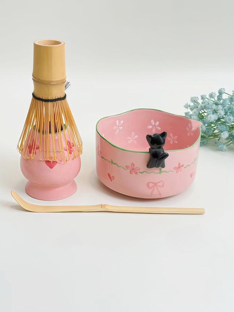 May include: A pink ceramic matcha bowl with a black cat figurine on the rim, a bamboo whisk on a matching pink stand, and a long bamboo scoop. The bowl features painted pink flowers, green vines, and small hearts on its exterior and interior.