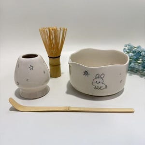 May include: A matcha tea set comprising a white ceramic bowl with a bunny illustration, a bamboo whisk, a bamboo scoop, and a ceramic whisk holder with star designs. The set is arranged on a white surface.