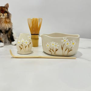 May include: A beige ceramic matcha tea set with a floral daisy design. The set includes a bowl, a whisk, a whisk holder, and a bamboo scoop. The bowl has a curved shape with a spout. The whisk is made of bamboo. The whisk holder is vase-shaped.