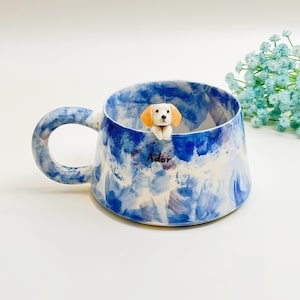 Può includere: Una tazza in ceramica con un motivo dipinto in stile acquerello blu e bianco. Una piccola figura di cucciolo bianco e marrone chiaro è fissata sul bordo interno. La parola Ador è stampata in nero sull'esterno. La tazza ha un manico largo e rotondo.