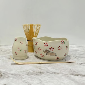 May include: A matcha tea set. The cream-coloured bowl has a dachshund illustration and red floral accents. Includes a whisk, a small vase, and a bamboo scoop. The set is designed for preparing matcha tea.