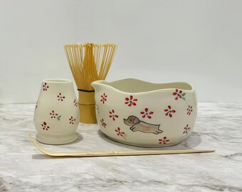 May include: A matcha tea set featuring a cream-colored bowl with a dachshund illustration and red floral accents. Includes a whisk, a small vase, and a bamboo scoop. The set is designed for preparing matcha tea.