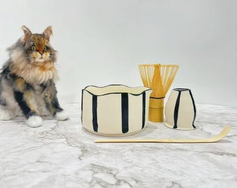 May include: A ceramic matcha bowl with a wavy rim and black vertical stripes, a bamboo whisk, a small matching ceramic cup with black stripes, and a long, curved bamboo tea scoop, all arranged next to a fluffy, multi-colored cat figurine.