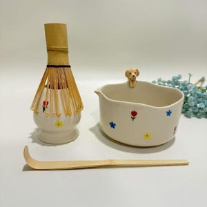 Könnte beinhalten: Ein Matcha-Teeset mit einem Bambusbesen, einer Keramikschale mit einer Hundefigur und einem Bambuslöffel. Die Schale und der Quirlhalter sind mit bunten Blumenmustern verziert. Das Set ist für die Zubereitung von Matcha-Tee konzipiert.
