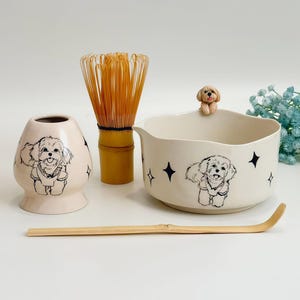 Op de afbeelding: Een matcha theeset met een crèmekleurige keramische kom en gardehouder, beide versierd met zwarte lijntekeningen van een kleine hond en sterren. De set bevat een bamboe garde en een bamboe lepel, met een klein hondenfiguurtje op de rand.