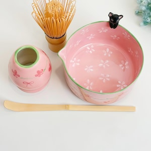 May include: A matcha tea set featuring a pink ceramic bowl with white cherry blossom patterns and a small black cat figurine on the rim, a pink whisk holder, a bamboo whisk, and a long, thin bamboo tea scoop for preparing traditional tea.