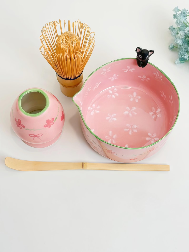 May include: A matcha tea set featuring a pink ceramic bowl with white cherry blossom patterns and a small black cat figurine on the rim, a pink whisk holder, a bamboo whisk, and a long, thin bamboo tea scoop for preparing traditional tea.