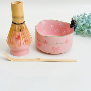May include: A pink ceramic matcha bowl with painted flowers and a small black cat figure on the rim, accompanied by a bamboo whisk on a pink stand and a long, curved bamboo tea scoop. The bowl features a green rim and a pink ribbon design.