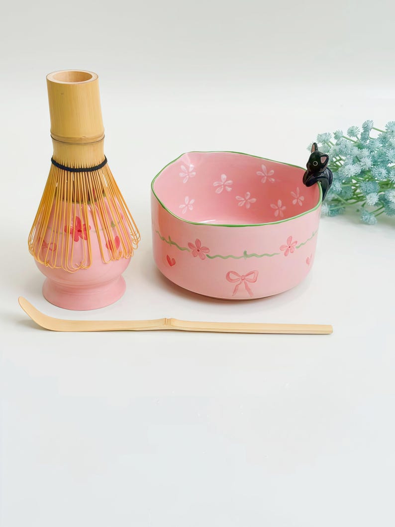 May include: A pink ceramic matcha bowl with painted flowers and a small black cat figure on the rim, accompanied by a bamboo whisk on a pink stand and a long, curved bamboo tea scoop. The bowl features a green rim and a pink ribbon design.