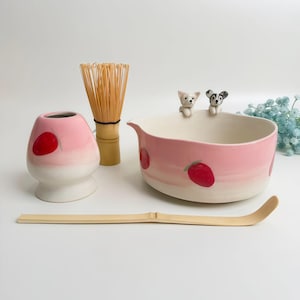 May include: A matcha tea set featuring a pink and white ceramic bowl with strawberry designs and two small dog figurines. Includes a bamboo whisk, a pink and white ceramic container with a red dot, and a bamboo scoop.