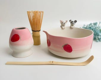 May include: A matcha tea set featuring a pink and white ceramic bowl with strawberry designs and two small dog figurines. Includes a bamboo whisk, a pink and white ceramic container with a red dot, and a bamboo scoop.