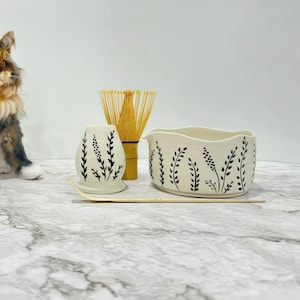 May include: A matcha tea set featuring a white ceramic bowl and a matching whisk holder, both adorned with black leaf patterns. A bamboo whisk and a wooden scoop are also included. The set is designed for preparing matcha tea.