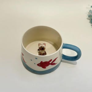 May include: A white ceramic mug with a blue handle and a blue band around the base. The mug features a 3D cat figurine inside and red fish illustrations on the exterior. The mug is designed for beverages.