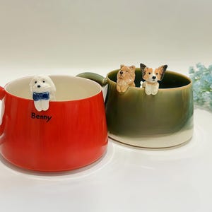 May include: A bright red ceramic mug with the name Benny printed on the front and a small white dog figurine with a blue bow attached to the rim, next to an olive green mug featuring two small dog figurines perched on the inner rim.