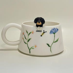 Può includere: Tazza in ceramica bianca con una statuina di cane nero e marrone sul bordo. La tazza è decorata con illustrazioni floreali e di api, e il nome "annie" è scritto sul lato. Il manico ha una forma arrotondata e curva.