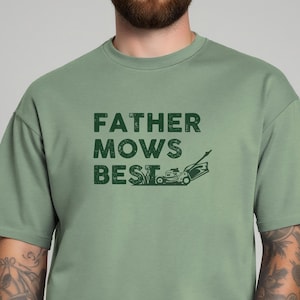 Peut inclure: T-shirt vert sauge avec l'inscription "FATHER MOWS BEST" dans une police de caractères vieillie, accompagnée d'un dessin de tondeuse à gazon. Le t-shirt a des manches courtes et un col rond.