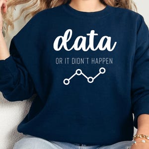 Könnte beinhalten: Marineblaues Sweatshirt mit dem weißen Text "data OR IT DIDN'T HAPPEN" und einem weißen Liniendiagramm. Das langärmlige Sweatshirt mit Rundhalsausschnitt wird mit blauen Jeans und goldenen Armbändern getragen.